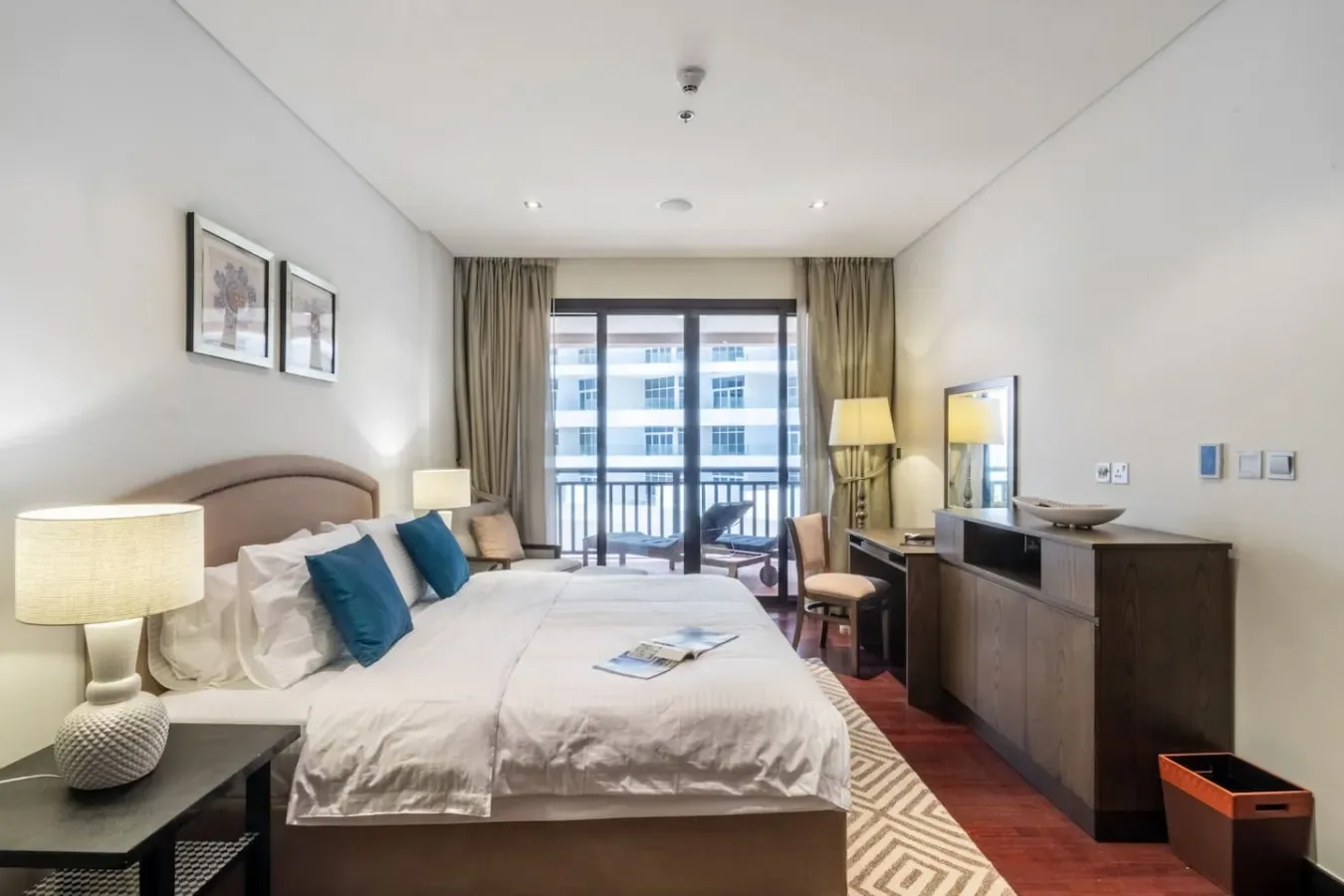 Un dormitorio con una cama grande, lámparas de noche y una ventana con vista a edificios cercanos.