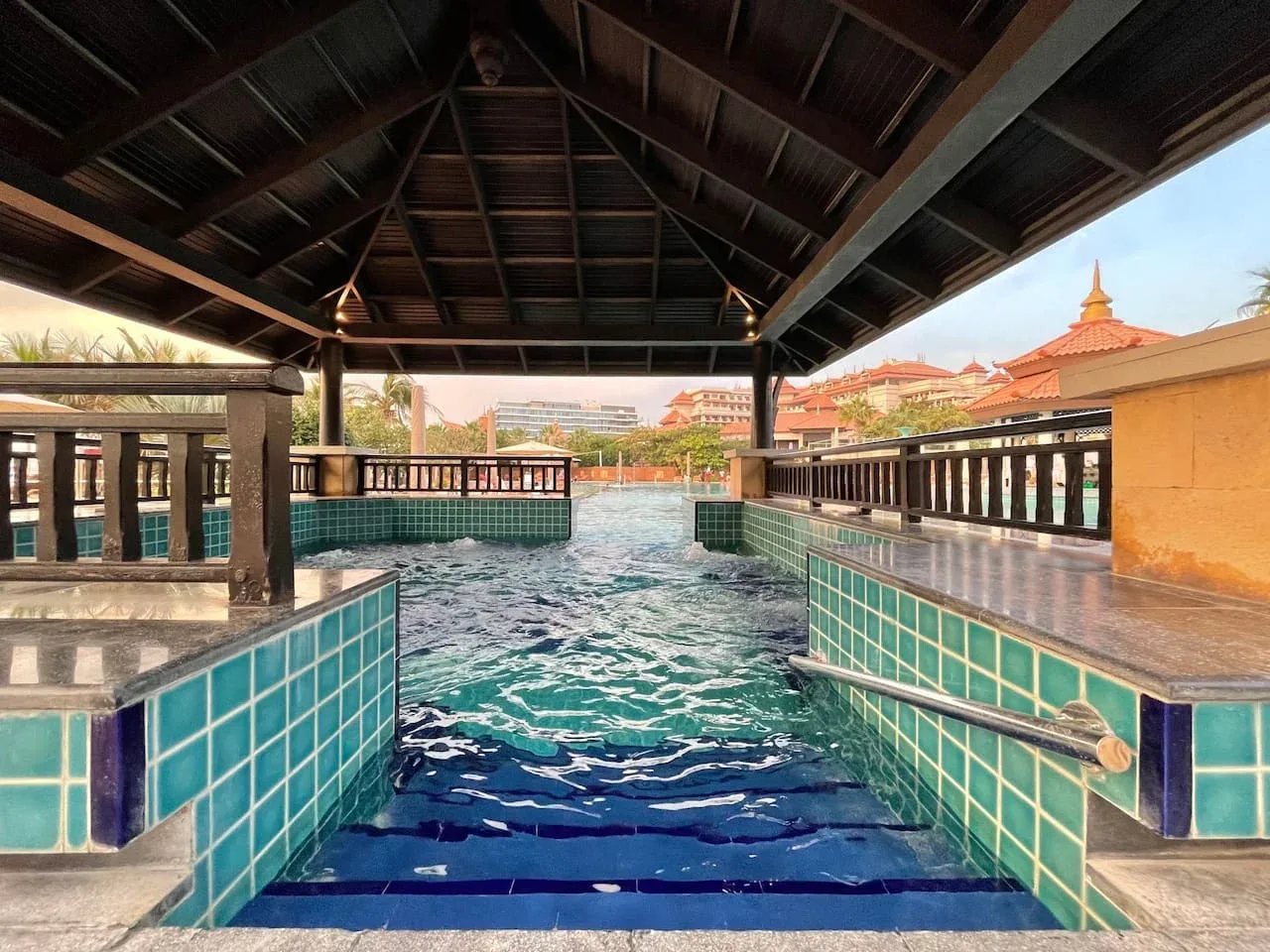 Una piscina revestida de azulejos está bajo un techo de pabellón de madera, con edificios al fondo.