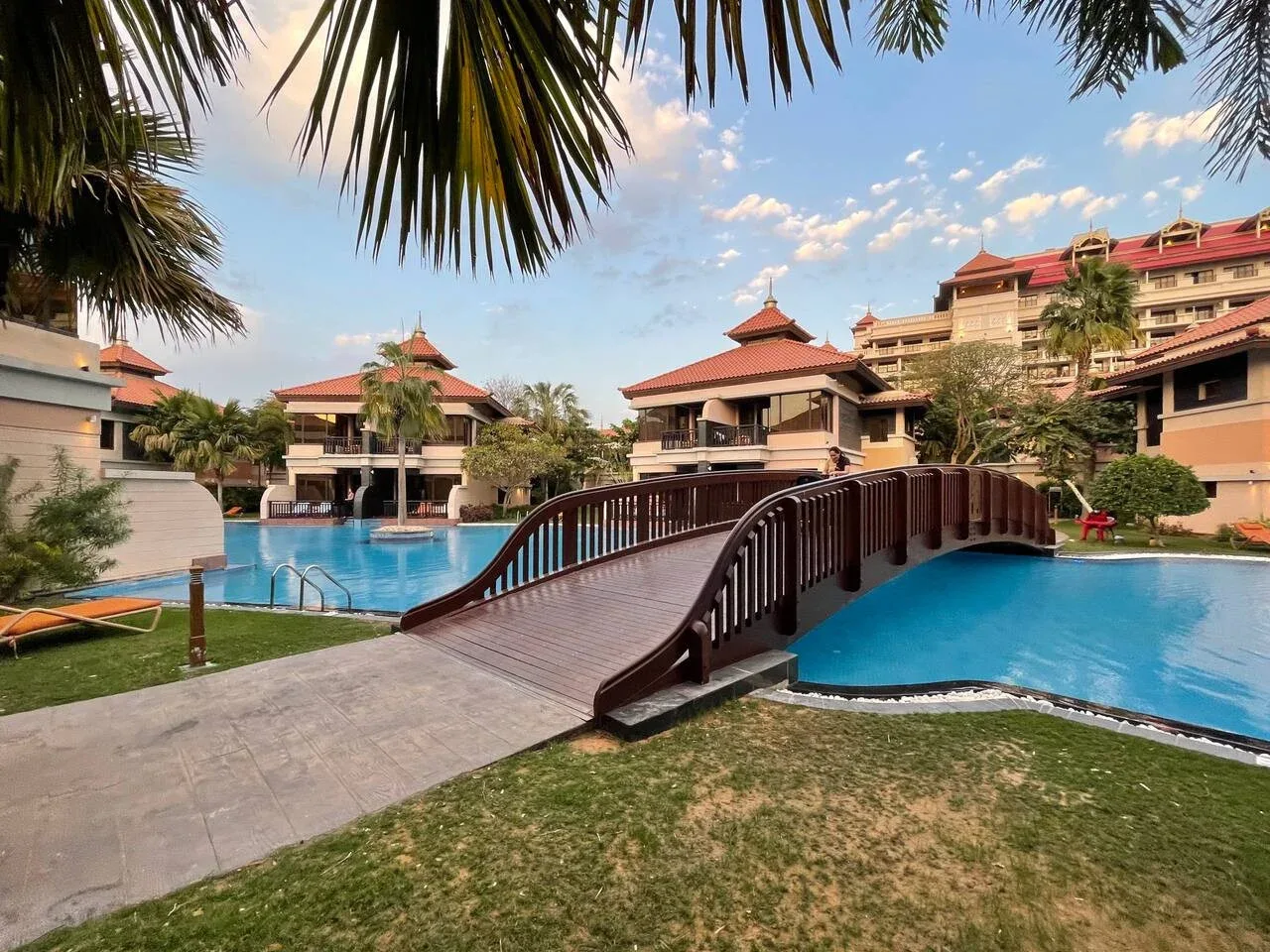 Un puente curvo de madera cruza una piscina entre edificios de estilo tropical.