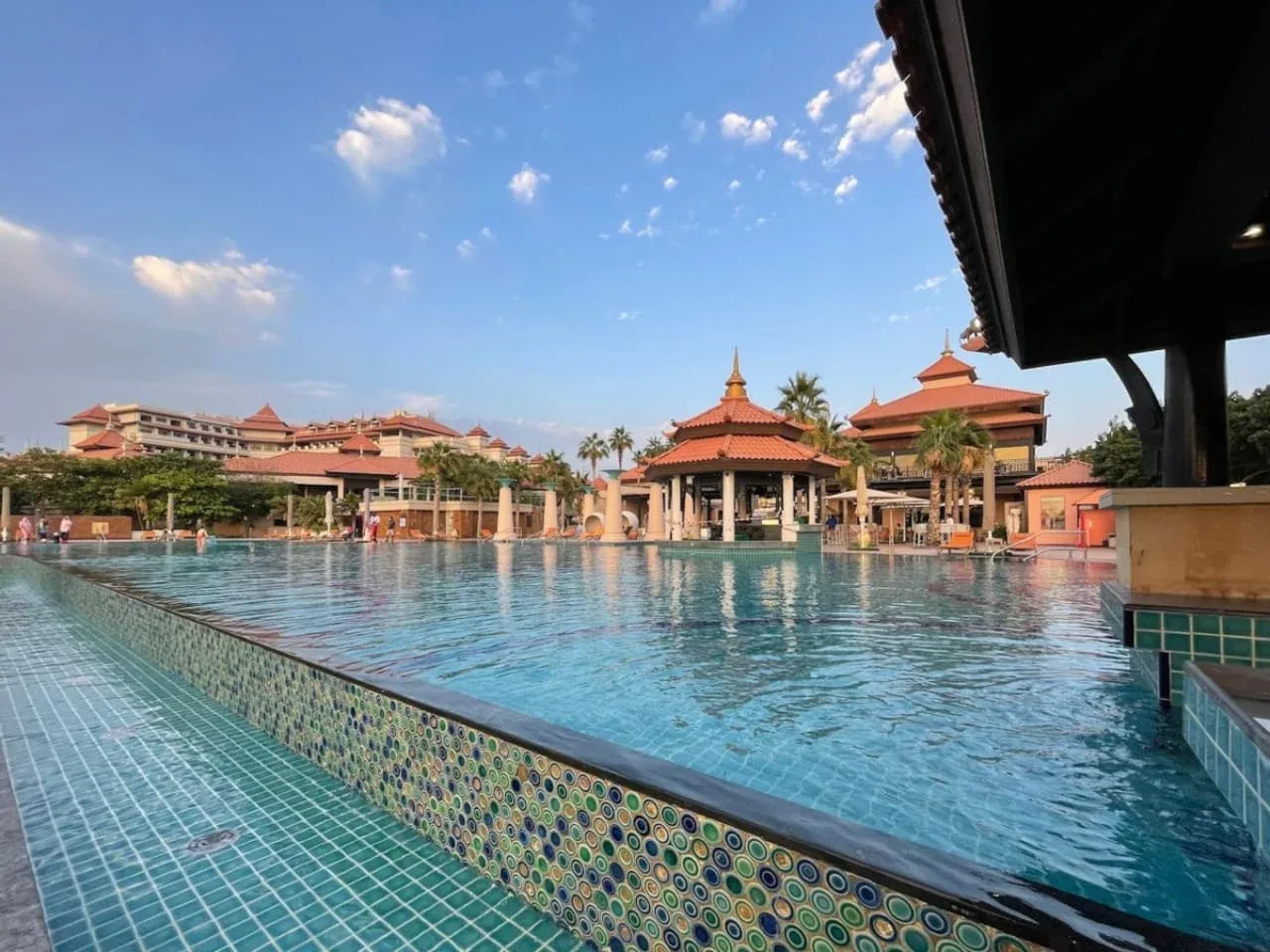 Una gran piscina al aire libre se encuentra frente a edificios de tejados rojos bajo un cielo azul.