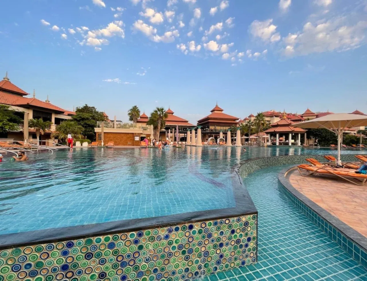 Una piscina de complejo turístico con bordes de azulejos, tumbonas y edificios bajos bajo un cielo azul.