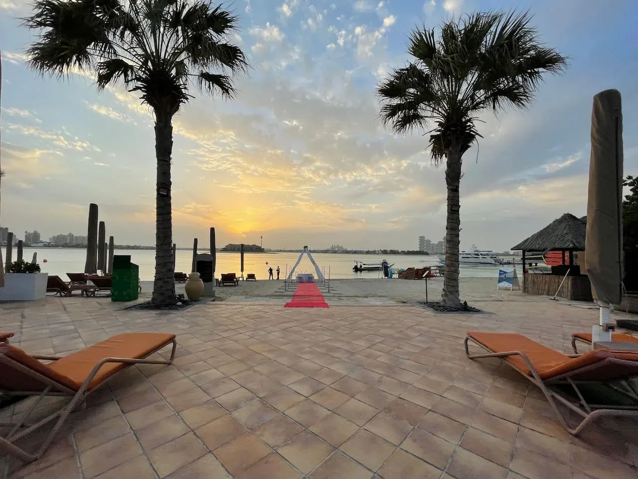 Una terraza embaldosada frente al agua, con asientos y palmeras, da a la playa al atardecer.