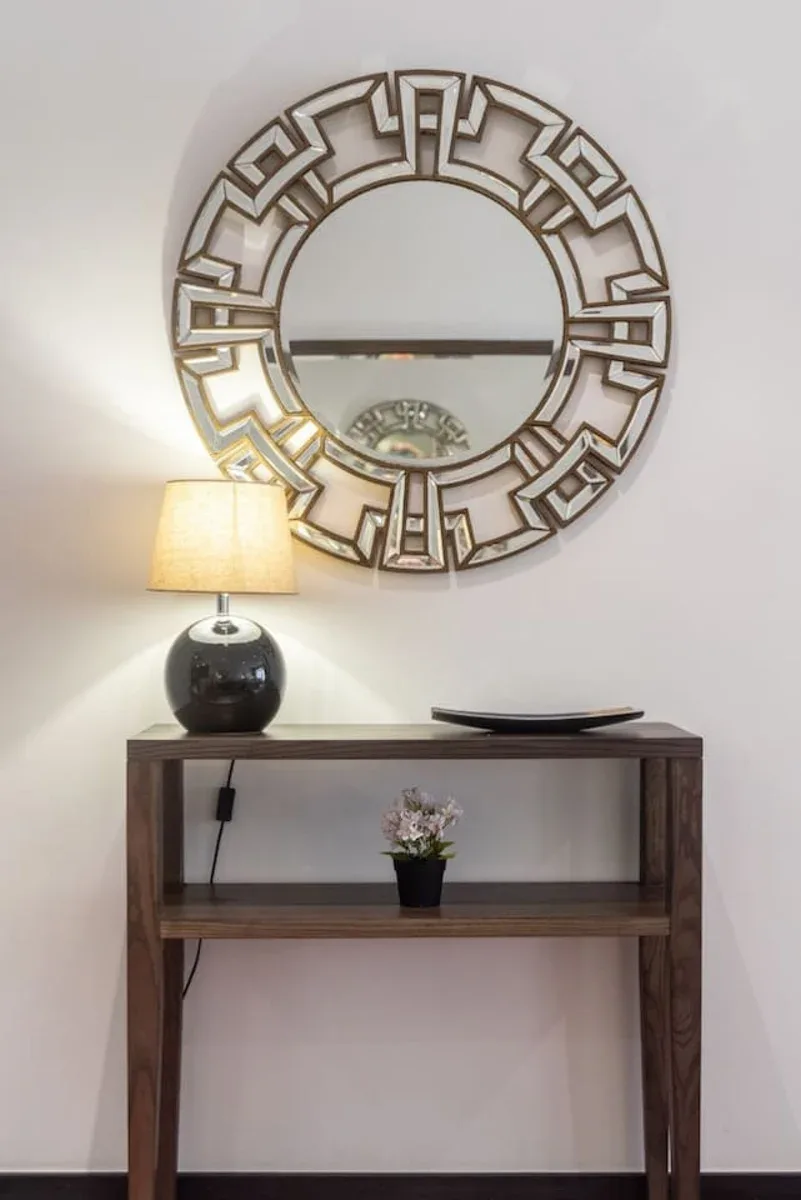 Una mesa consola en un pasillo está debajo de un gran espejo decorativo redondo.