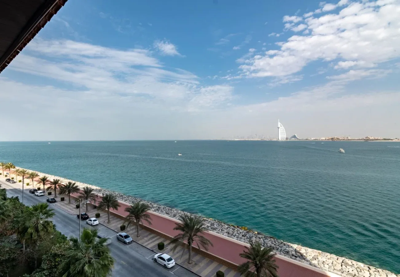 Una vista costera muestra el mar, una carretera bordeada de palmeras y una torre lejana con forma de vela.