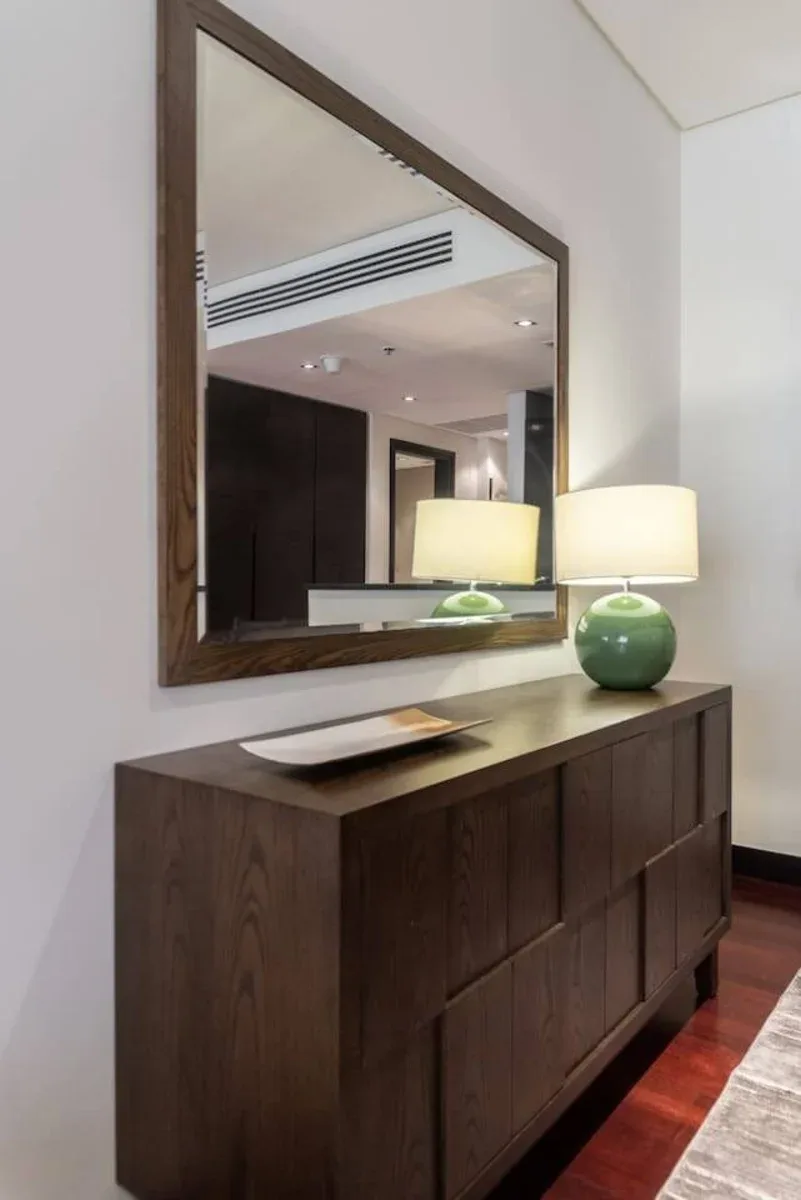 Una cómoda de madera oscura está bajo un espejo grande, junto a una lámpara encendida.