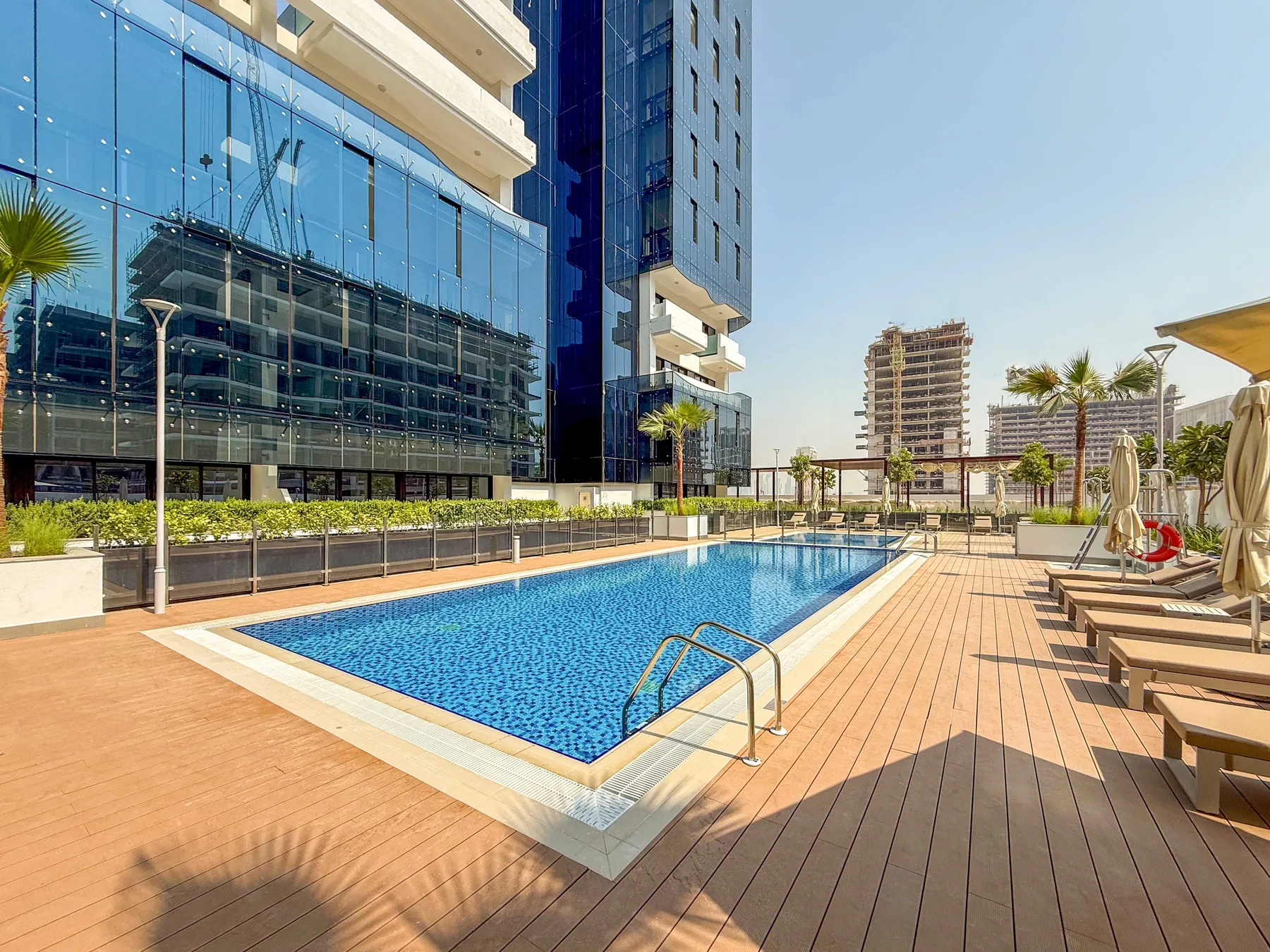 A swimming pool sits beside a sunlit wooden deck and modern buildings.