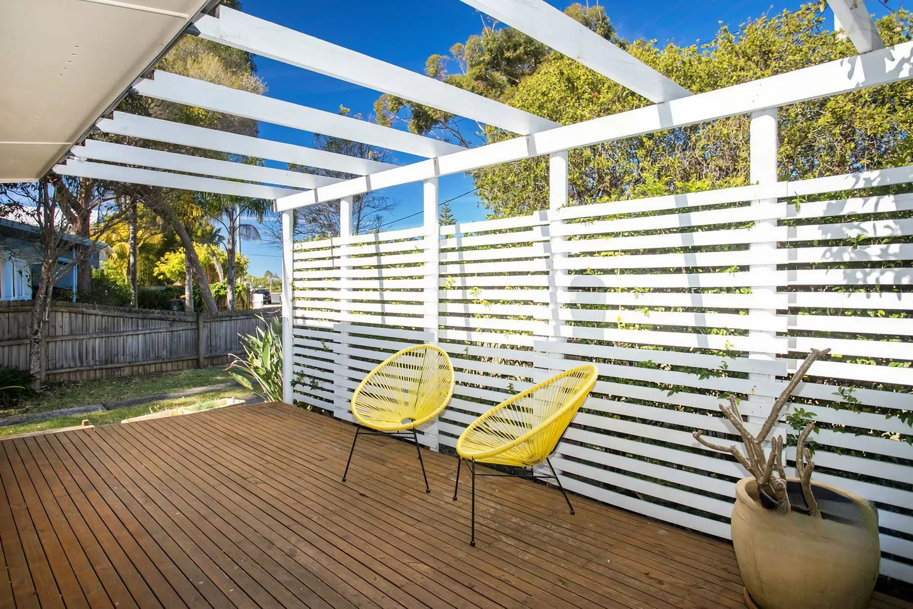 A wooden deck sits beneath a white pergola beside a slatted privacy screen.