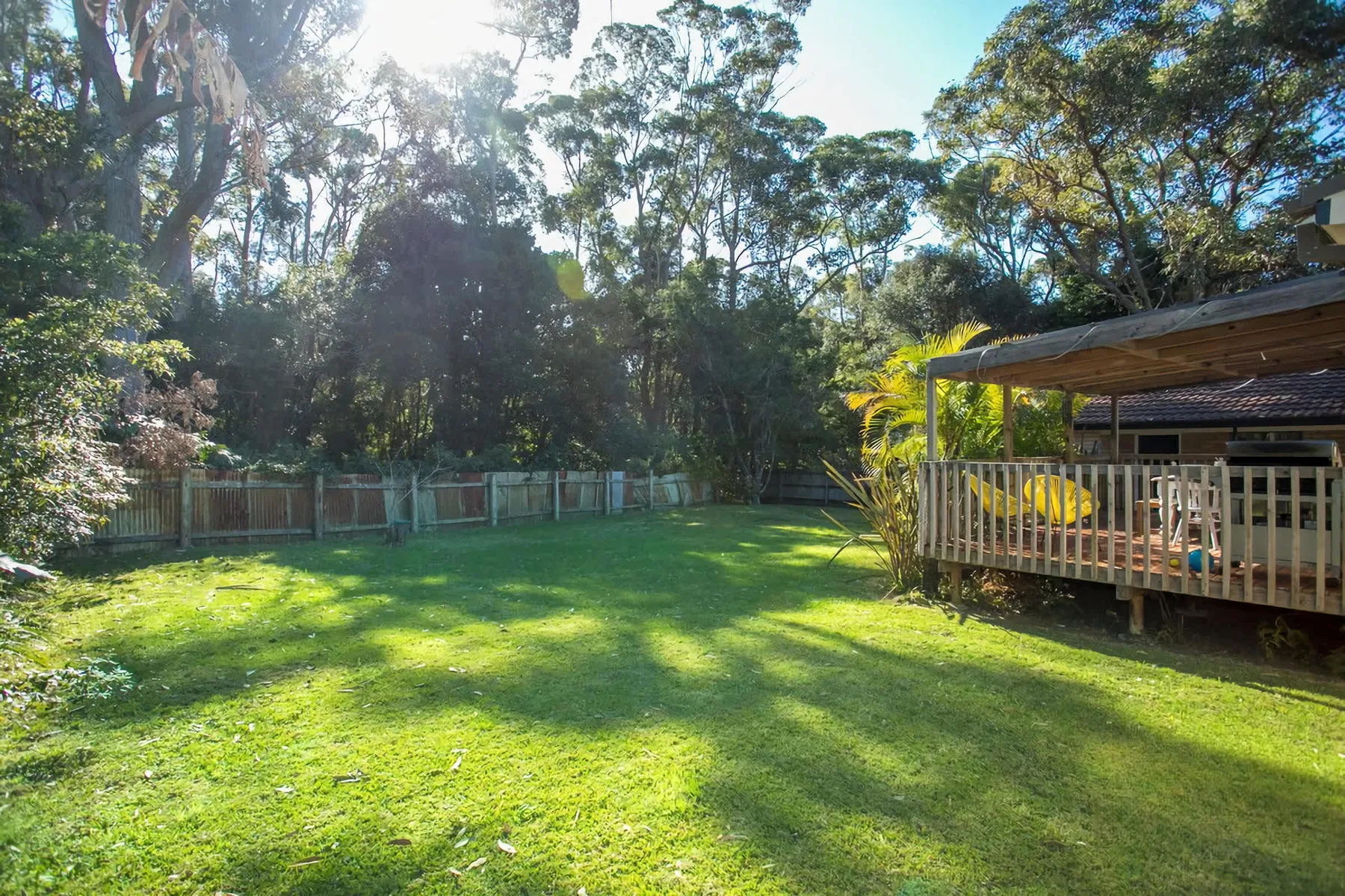 A sunny grassy yard bordered by trees, with a covered deck along one side.