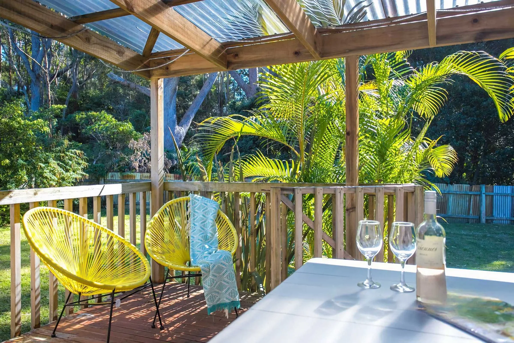 A covered wooden deck has two yellow seats and a table overlooking lush greenery.