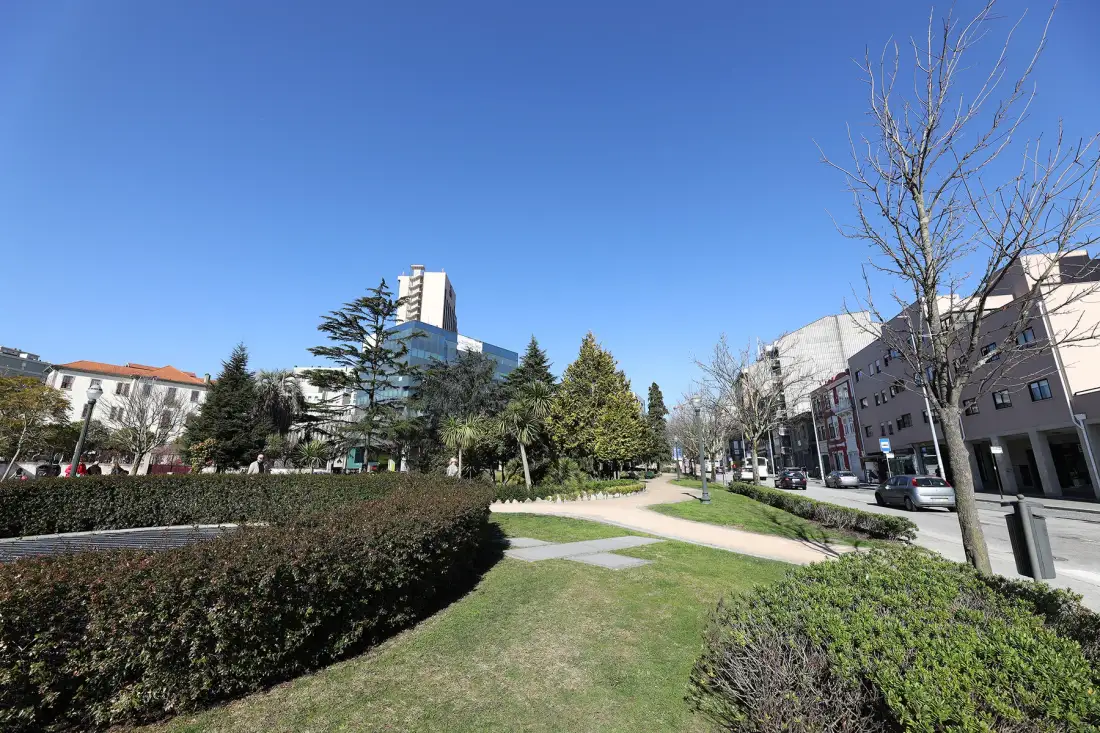 A landscaped outdoor area with a paved path, shrubs, trees, and surrounding buildings.