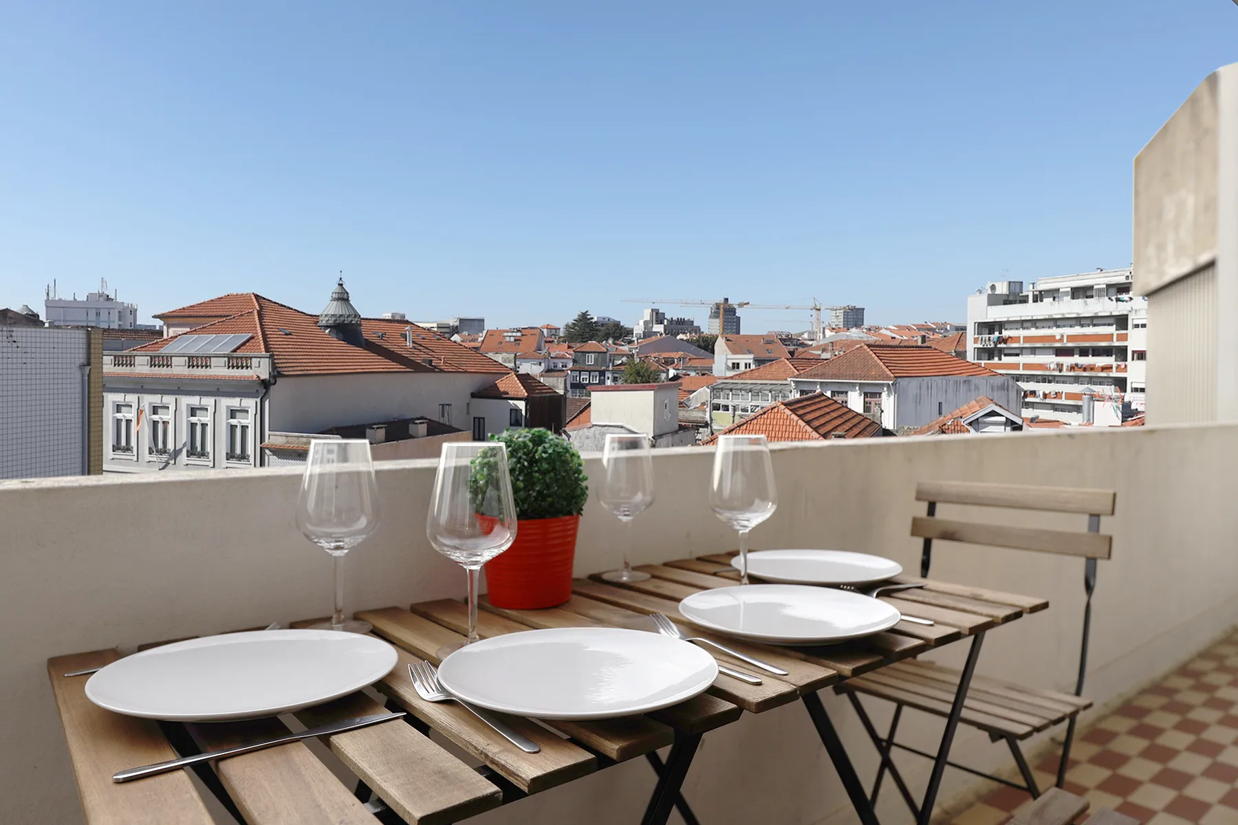 A rooftop terrace has a wooden dining table set with plates overlooking red-roofed buildings.