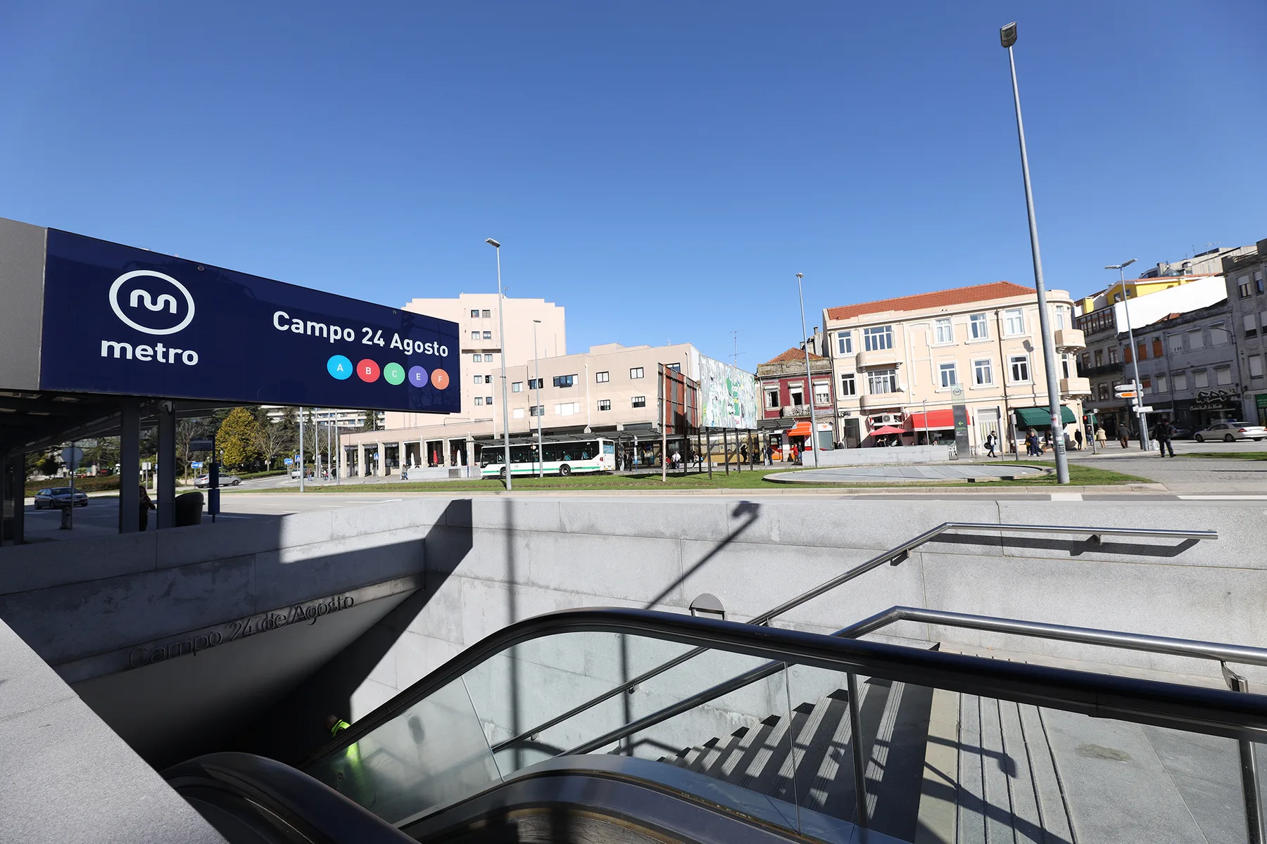 A metro station entrance opens onto a sunny urban plaza lined with buildings.