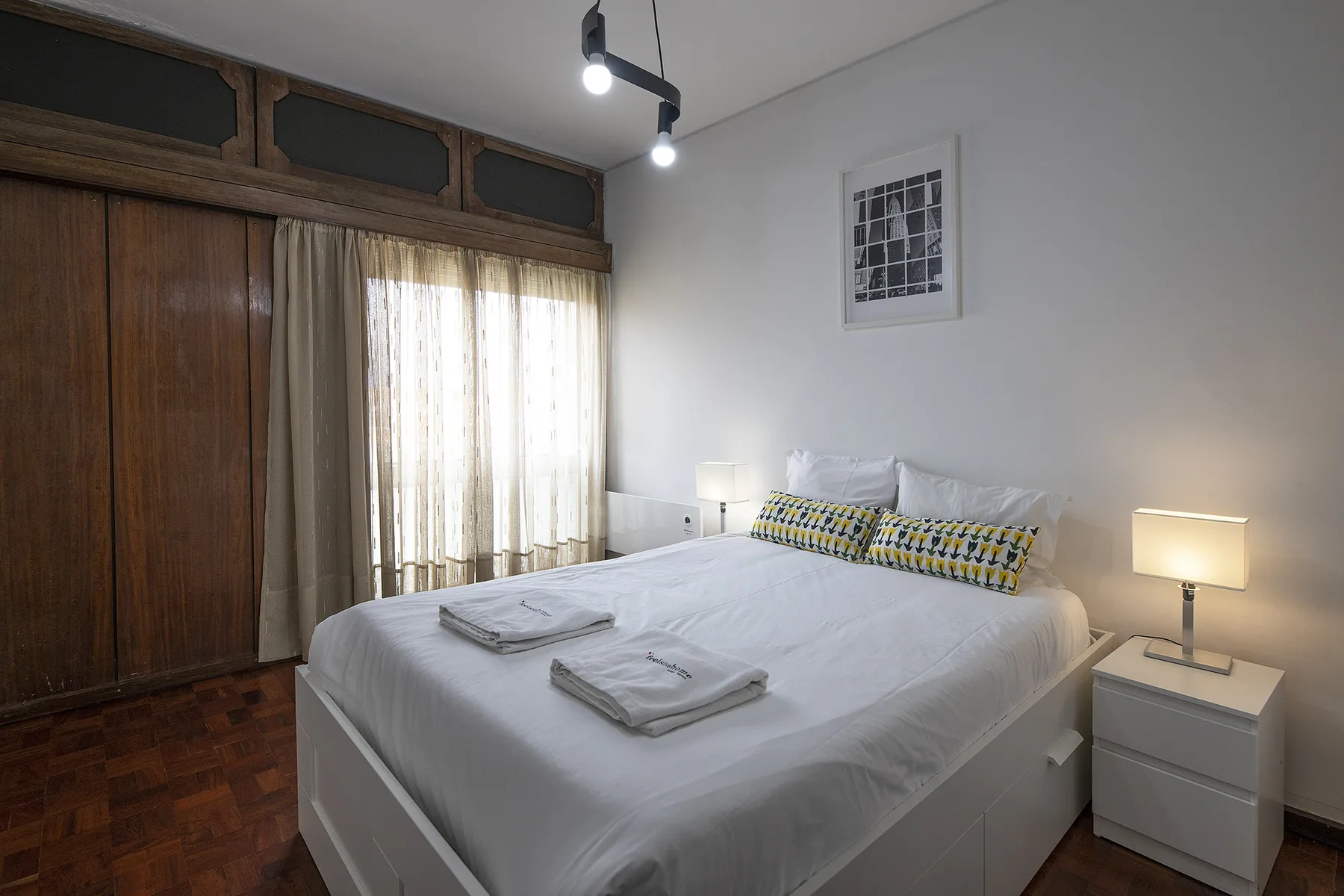 A bedroom with a neatly made bed, bedside tables, and a large wooden wardrobe.