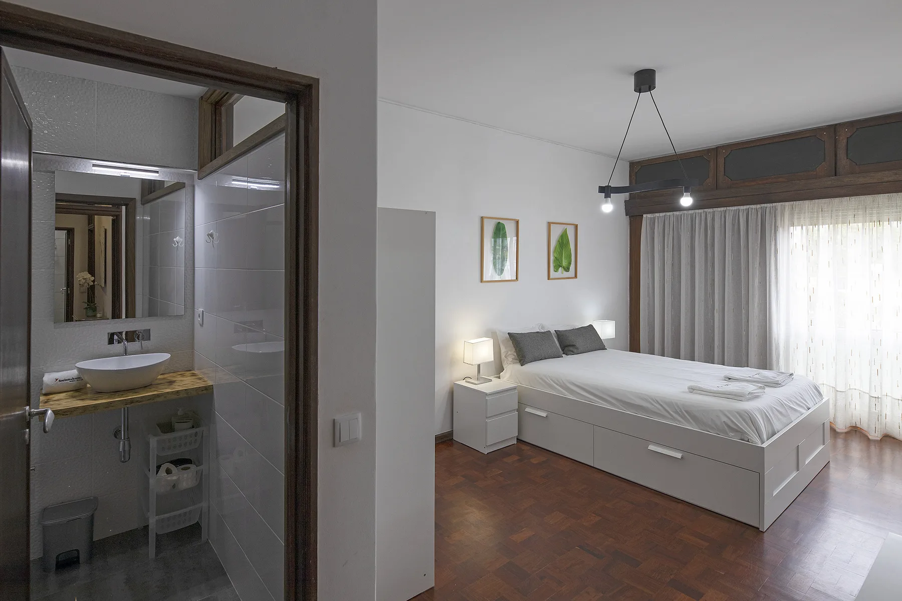 A bedroom with a bed beside a bathroom and a curtained window.