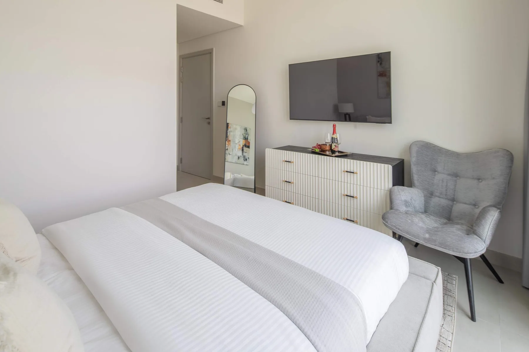 A bright bedroom with a neatly made bed, dresser, wall-mounted television, and armchair.