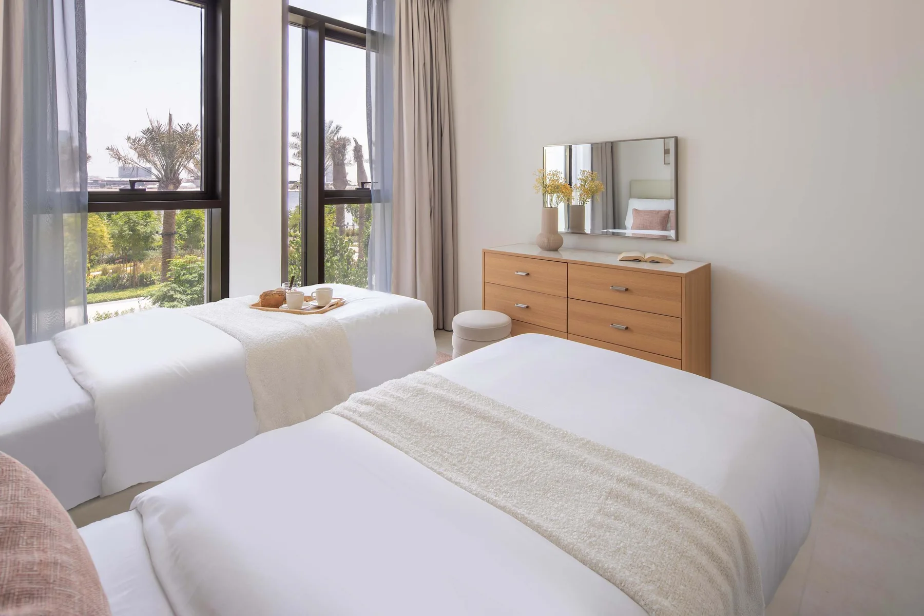 A bright bedroom with two beds, tall windows, and a wooden dresser.