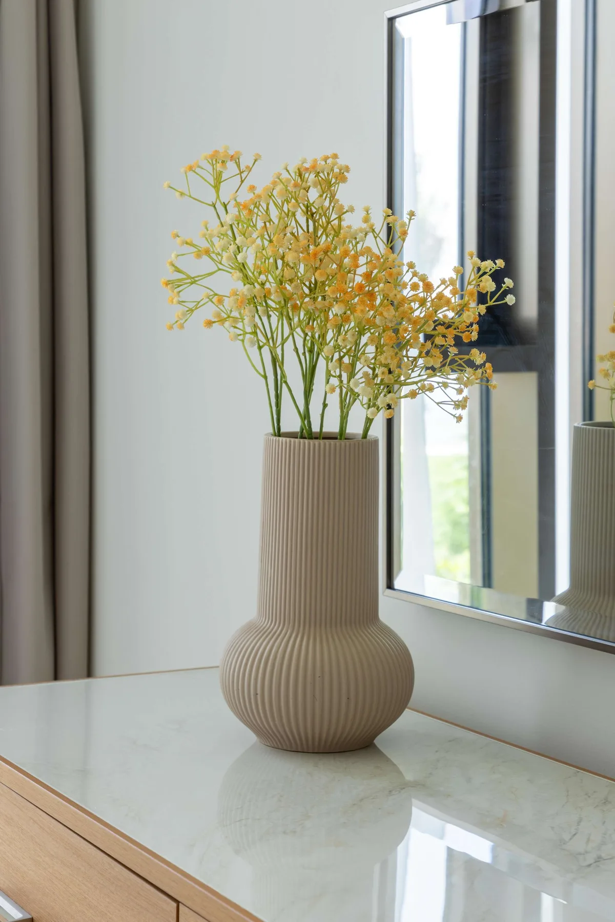 A ribbed vase of yellow flowers sits on a counter beside a mirror.