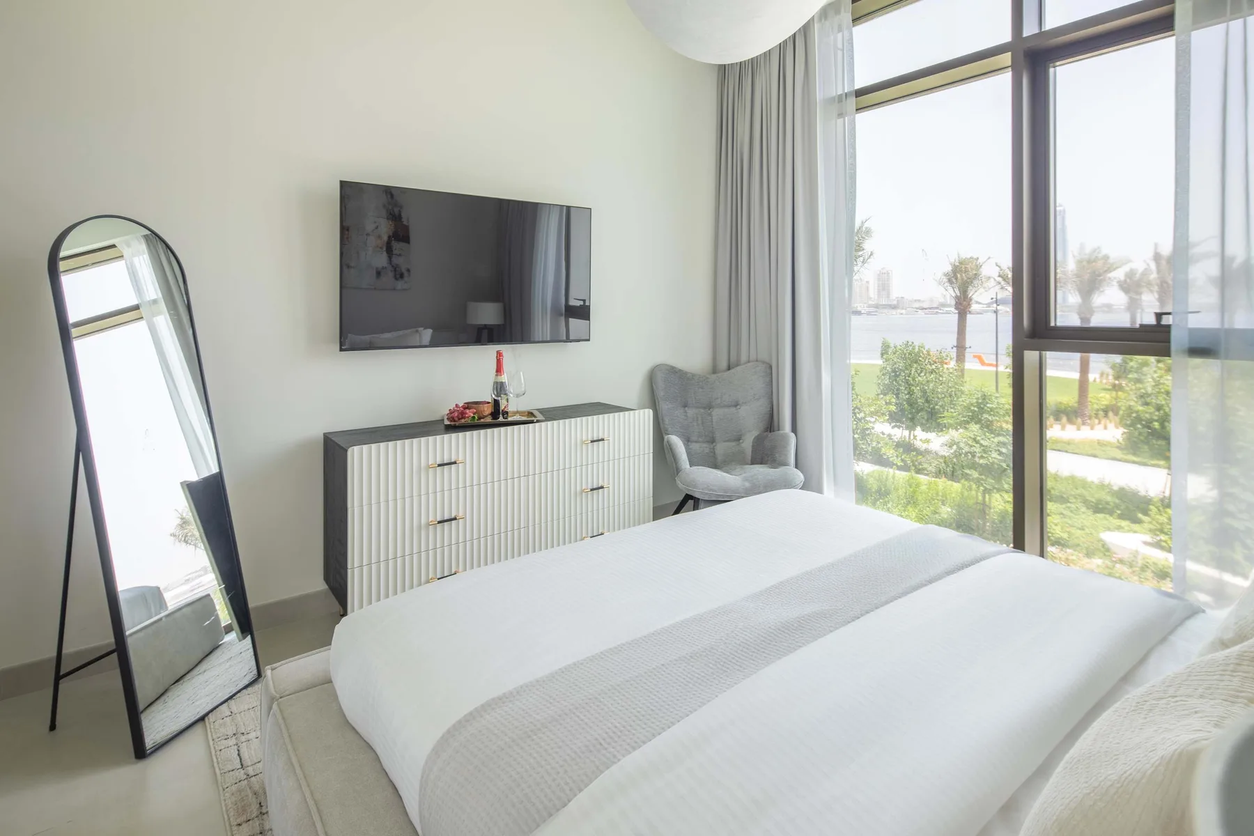 A bright bedroom with a neatly made bed, wall-mounted television, and large windows.