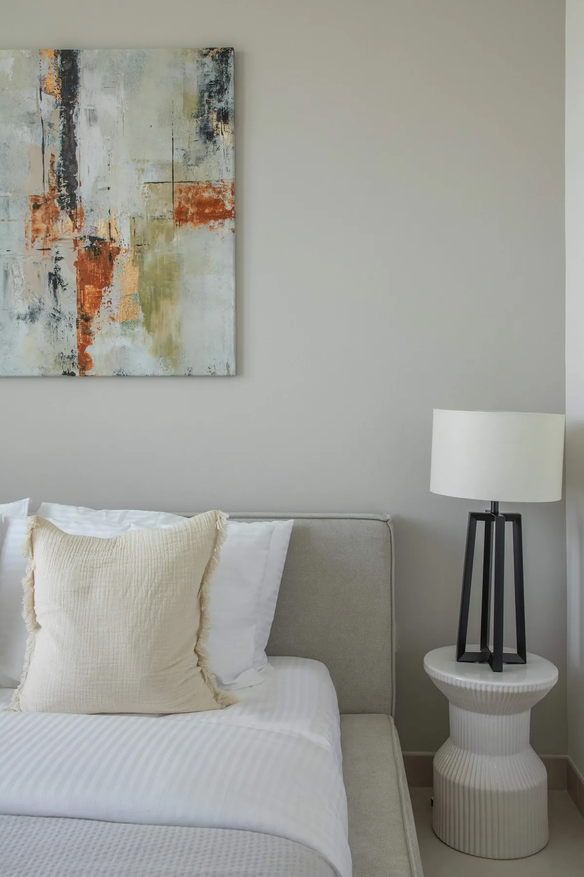 A neatly made bed sits beside a table lamp beneath abstract wall art.