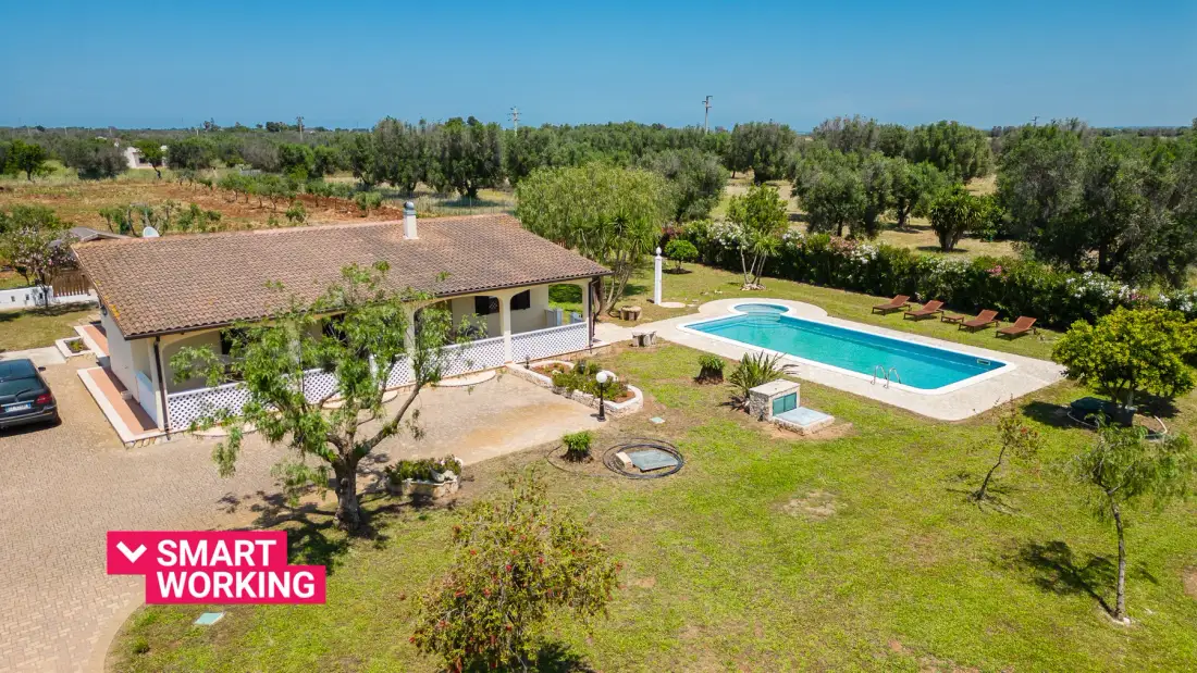 An outdoor swimming pool sits beside a tiled-roof building in a landscaped rural setting.