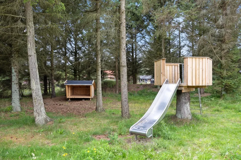 Un tobogán de madera y estructuras de juego están entre árboles en un claro cubierto de hierba.