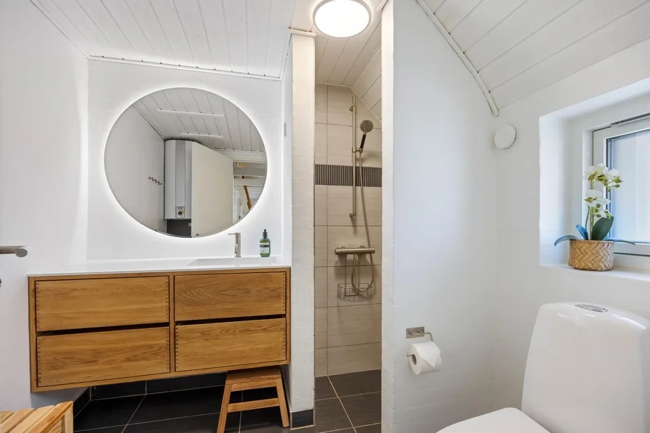 Un baño luminoso con un tocador de madera, espejo redondo y ducha.