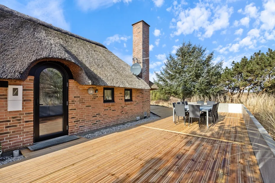 Una casa de ladrillo con techo de paja da a una terraza de madera con asientos al aire libre.