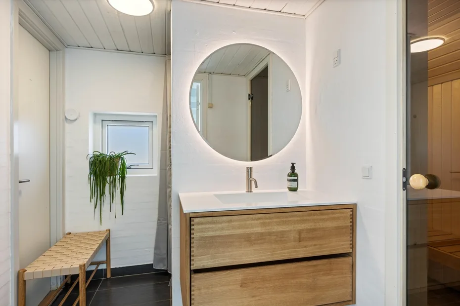Un baño luminoso con un mueble de madera, espejo redondo y una planta.