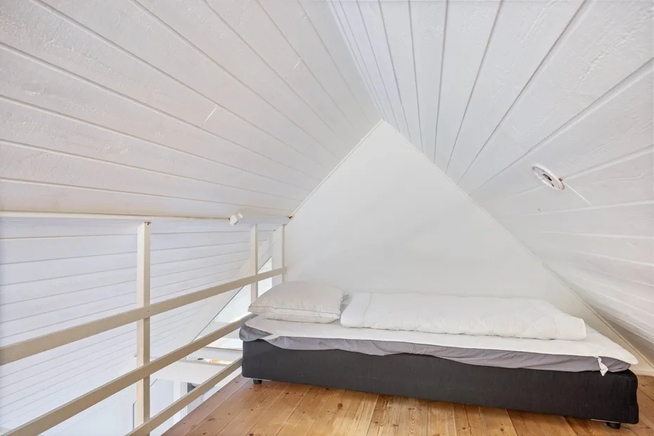 Una cama está bajo el techo blanco inclinado de un dormitorio tipo loft.