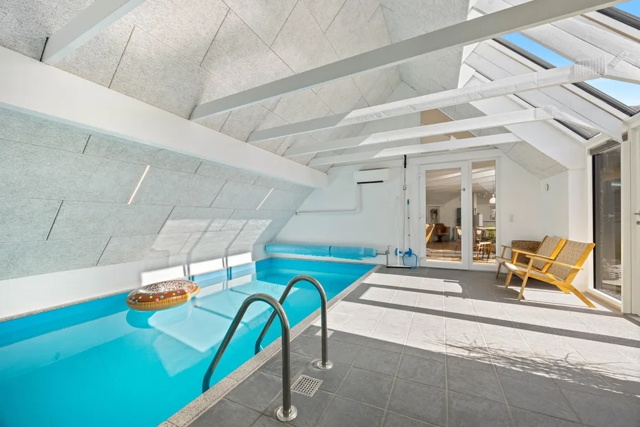 Una piscina interior luminosa bajo un techo inclinado de vidrio, con tumbonas cerca.