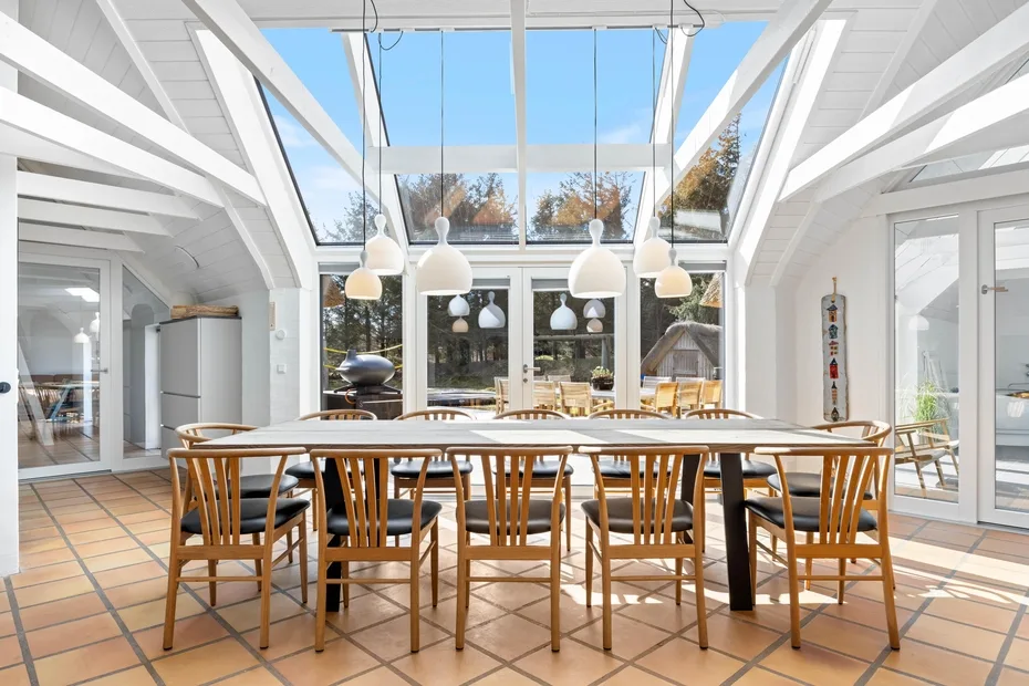 Un comedor luminoso tiene una mesa larga bajo grandes ventanas y luces colgantes.