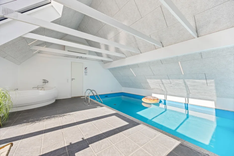 Una piscina cubierta luminosa bajo un techo inclinado, con una pequeña bañera elevada cerca.