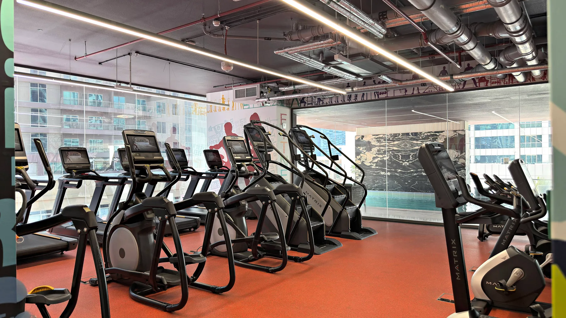 Un gimnasio con filas de máquinas de ejercicio y grandes ventanas.