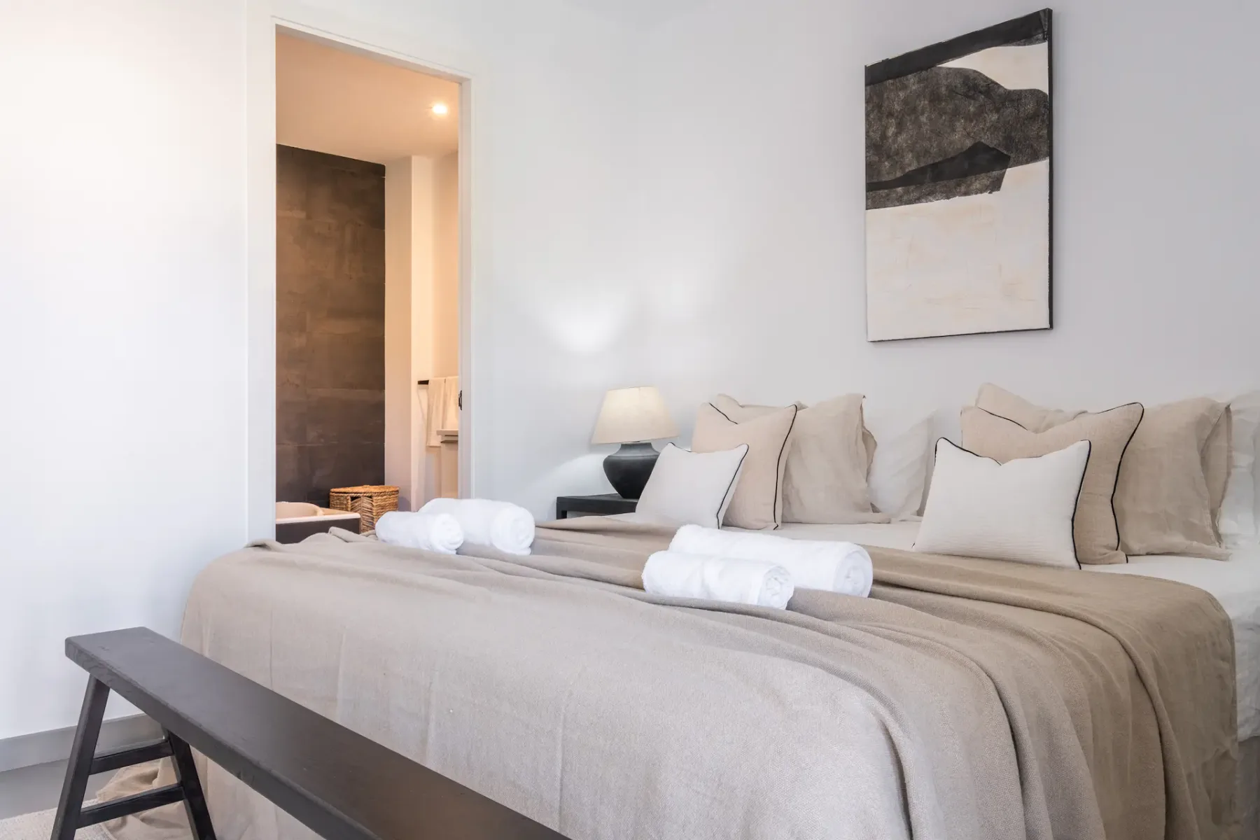 A neatly made bedroom with neutral bedding and an open doorway to a bathroom.