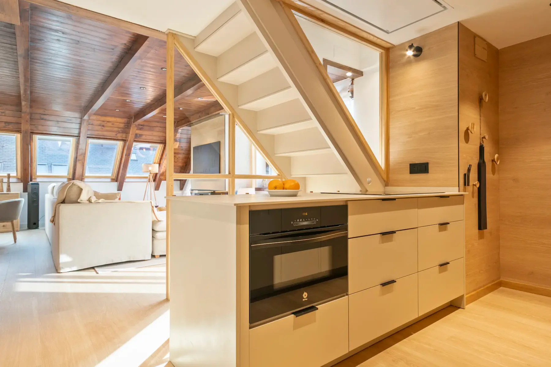 A modern kitchen and dining area with wooden furniture, a table set for four, pendant lights, and large windows allowing natural light. The room features light wood flooring and beige tones.