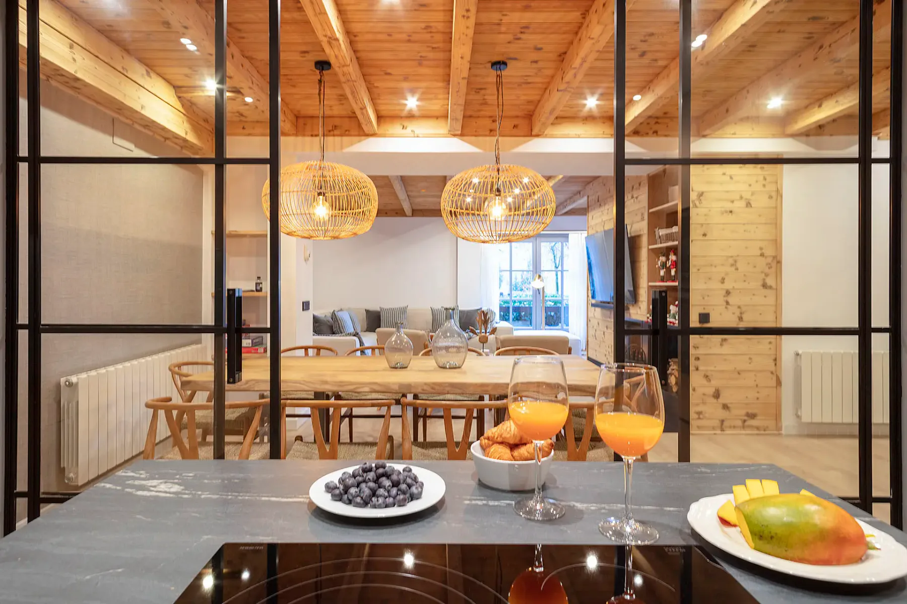 A modern kitchen and dining area with wooden furniture, a table set for four, pendant lights, and large windows allowing natural light. The room features light wood flooring and beige tones.