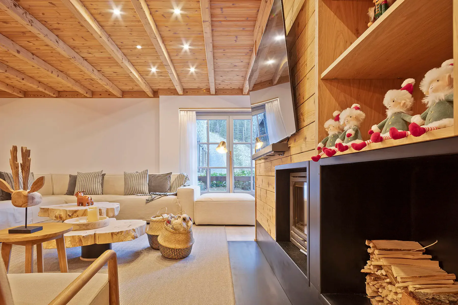 A modern open-concept living and dining area with wooden accents. A gray sofa, wooden coffee table, and poufs are in the foreground, while a dining table with chairs and kitchen are in the background. Large windows provide natural light.