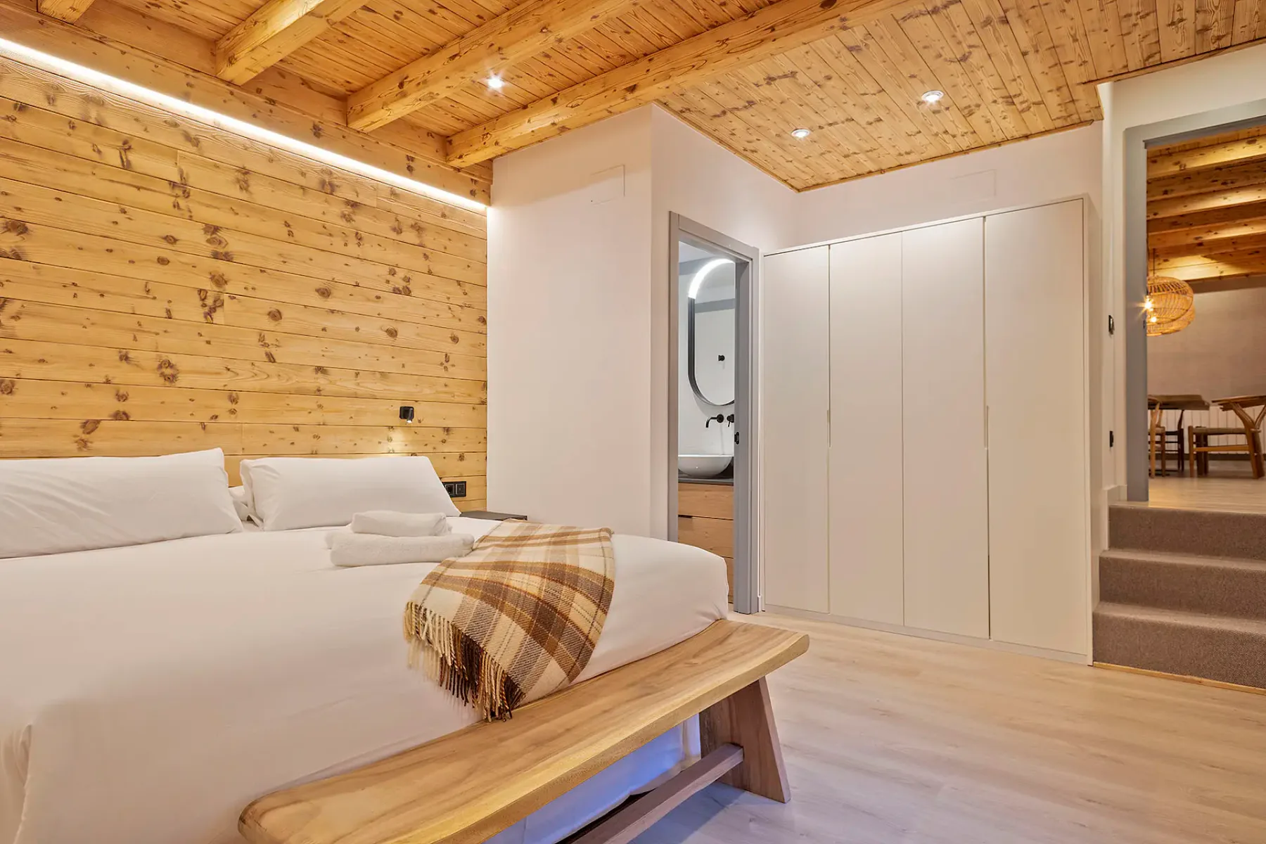 A cozy attic bedroom with a wooden ceiling and light blue walls. The room features a double bed with gray and white bedding, two lamps, a small wooden bench, and a skylight window.
