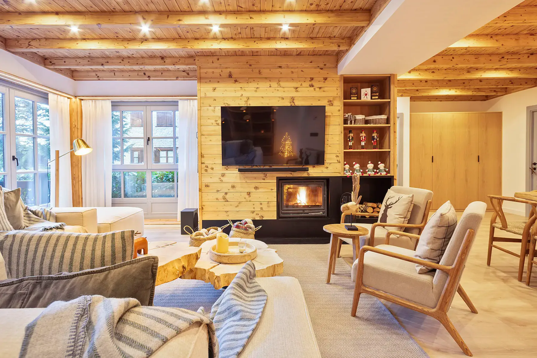 A modern living room with two gray sofas, a wooden coffee table, two round ottomans, large windows with mountain views, and a black fireplace.