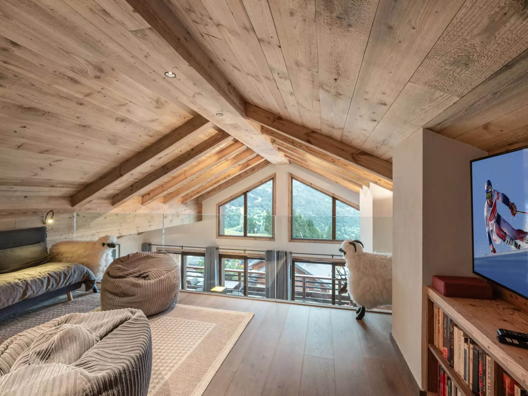 A loft lounge with seating, a bed, and large windows beneath a sloped wooden ceiling.