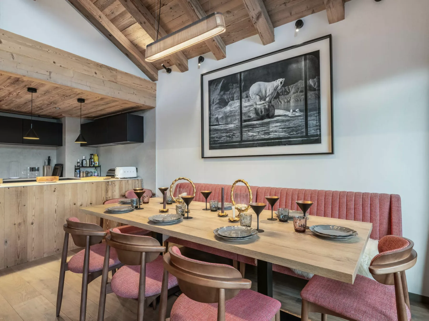 A dining area with a long wooden table, upholstered seating, and a compact kitchen.