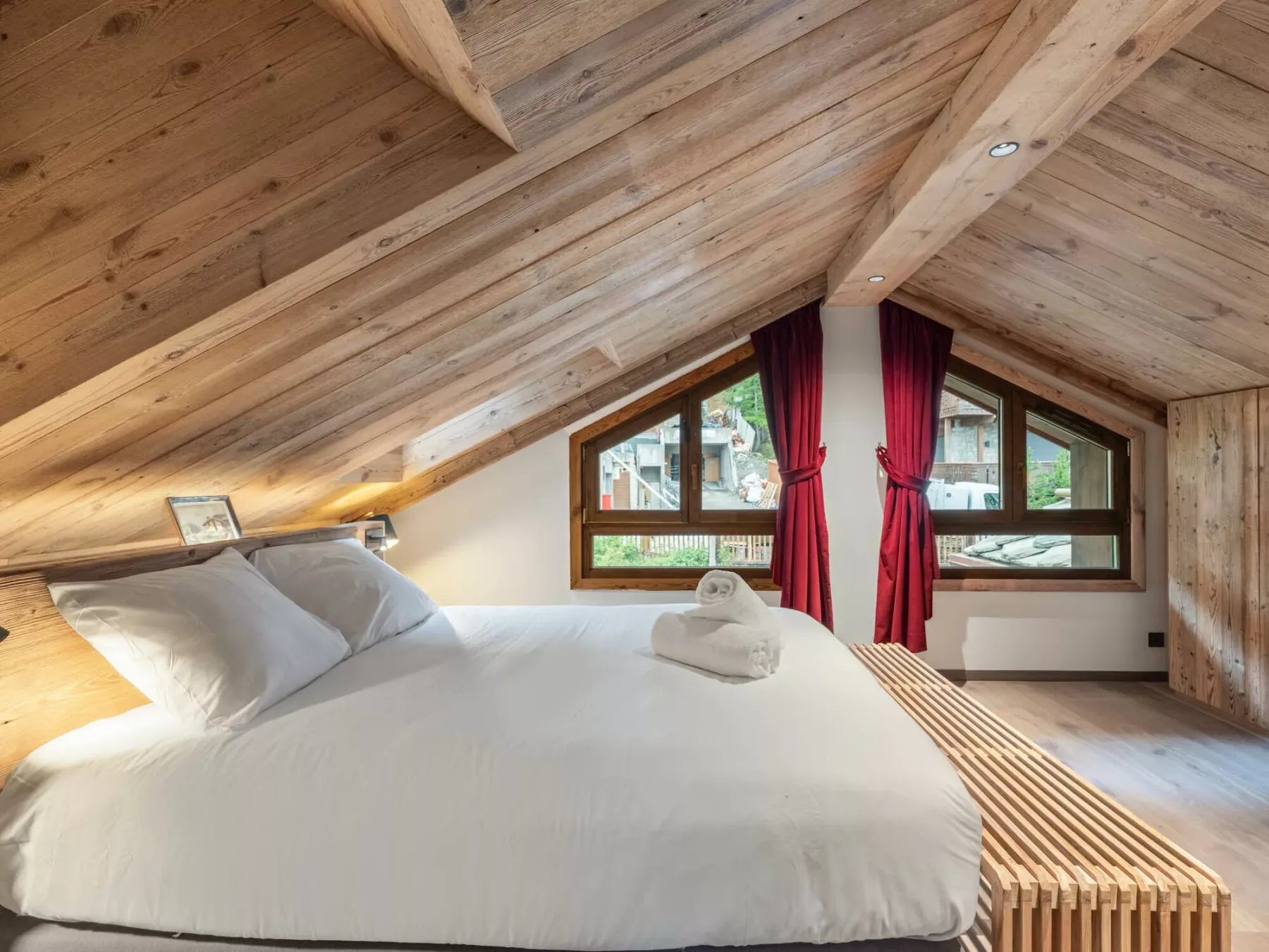 A bedroom with a bed beneath a sloped wooden ceiling and a window with red curtains.