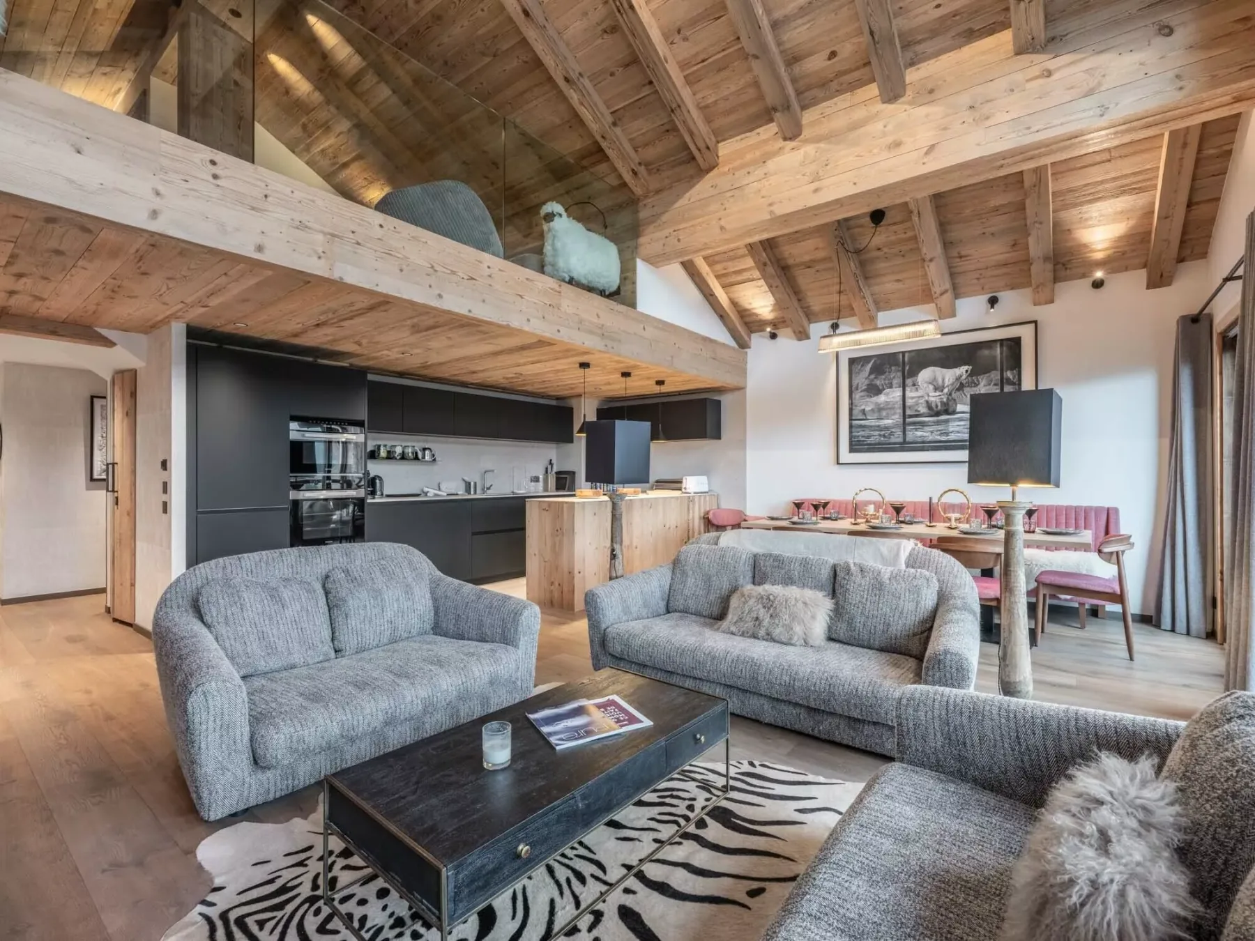 A living room with gray seating and a kitchen beneath a timber ceiling.