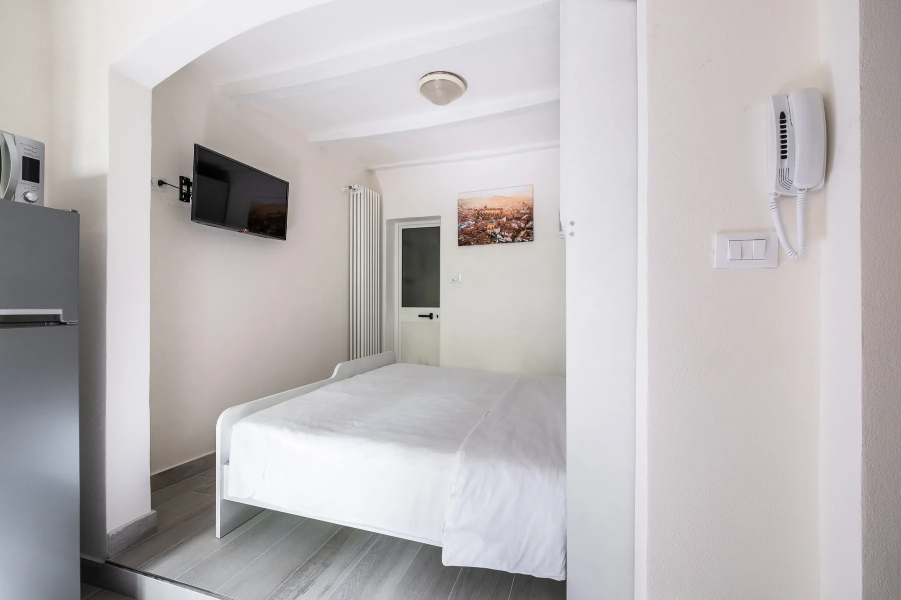 Un dormitorio con una cama blanca, un televisor en la pared y un refrigerador.