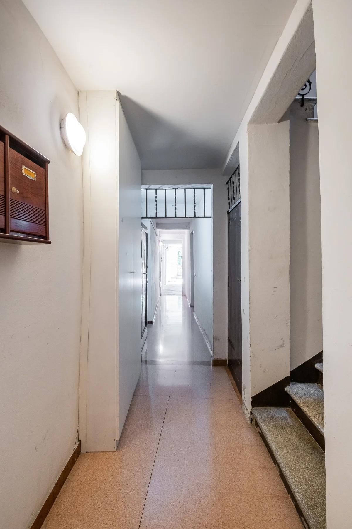 A narrow hallway leads toward a bright doorway, with a staircase along the side.