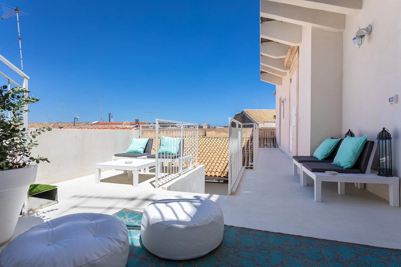 A sunny rooftop terrace has outdoor seating, floor cushions, and views of nearby rooftops.