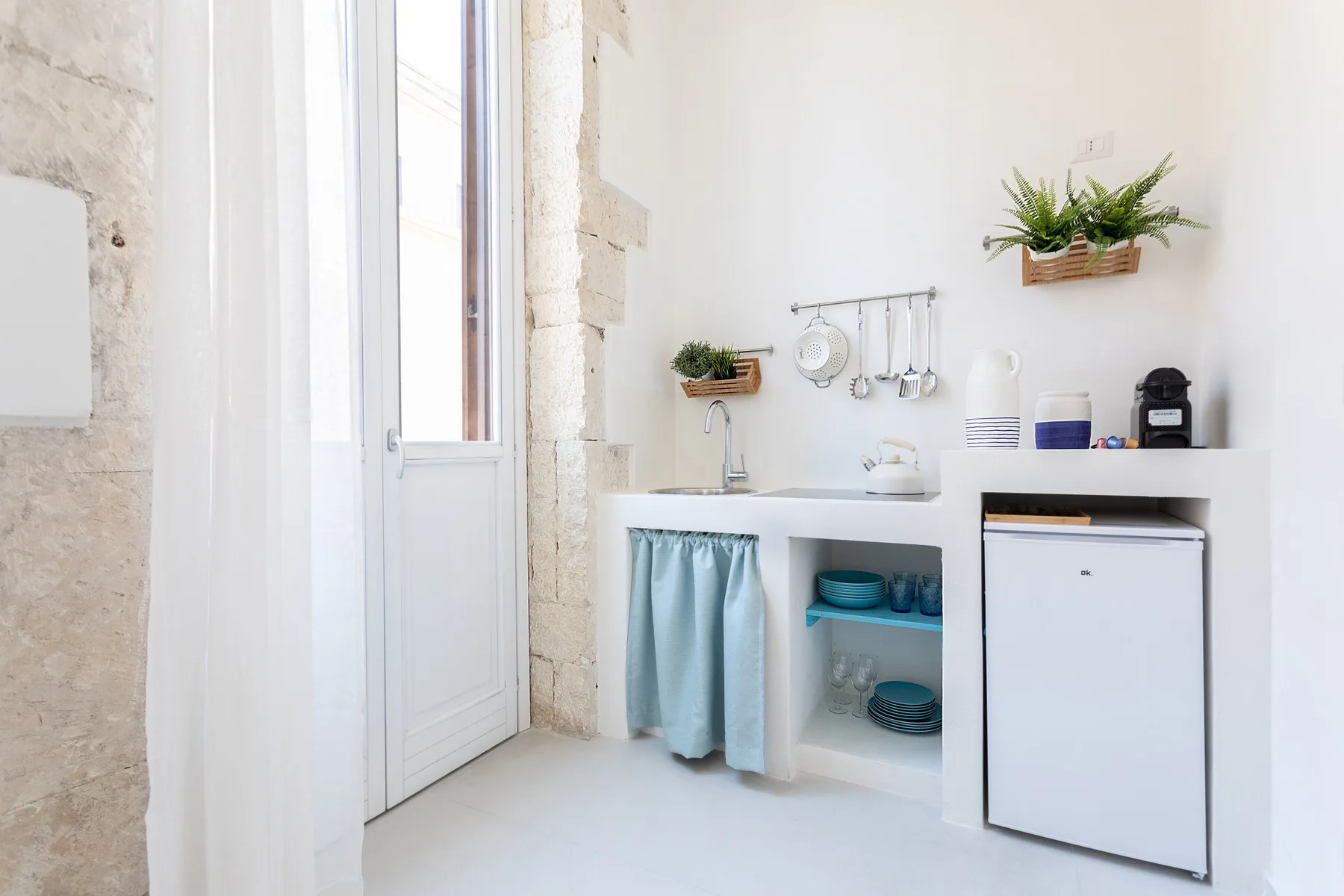 Una pequeña cocina luminosa con fregadero, estantes abiertos y refrigerador compacto.