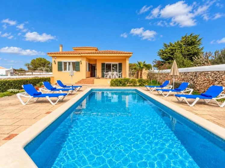 Una piscina exterior soleada junto a una casa de una planta, con tumbonas alrededor.