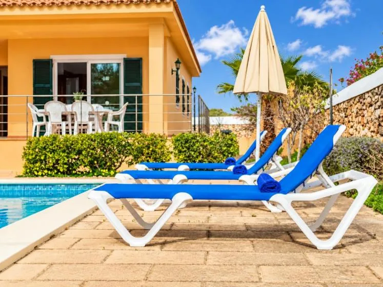 Un patio junto a la piscina con tumbonas azules y una sombrilla cerrada junto a una casa.