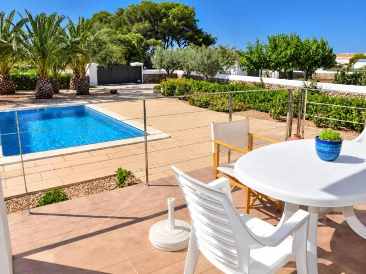 Una piscina exterior junto a un patio pavimentado con asientos blancos y una mesa.